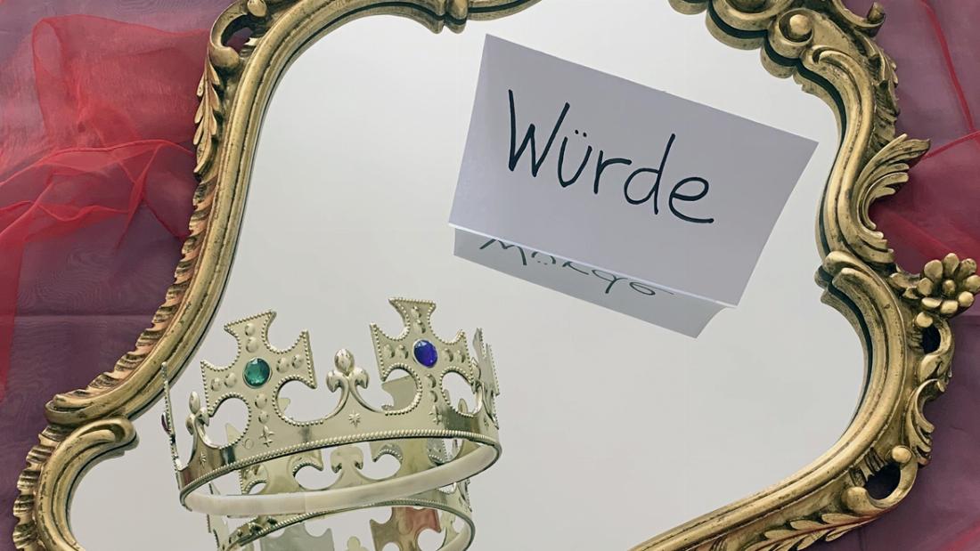Foto: Ein Spiegel liegt auf einem Seidentuch. Auf dem Spiegel liegen eine Krone und ein Zettel mit dem Wort "Würde".